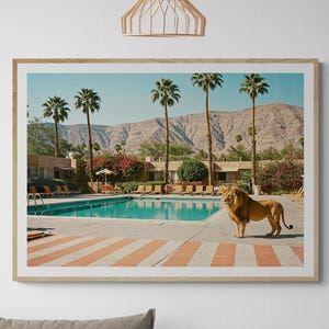 May include: Framed art print featuring a lion standing near a swimming pool with palm trees and mountains in the background. The pool has a turquoise water and is surrounded by lounge chairs. The artwork is displayed above a beige sofa.