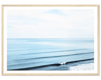 May include: A framed photograph of a seascape with a blue sky and ocean. The ocean is a light blue color and has a few small waves. The sky is a light blue color and has a few white clouds.