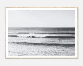 May include: A black and white photograph of ocean waves crashing on the shore. The waves are rolling in from the left side of the image and breaking into white foam.