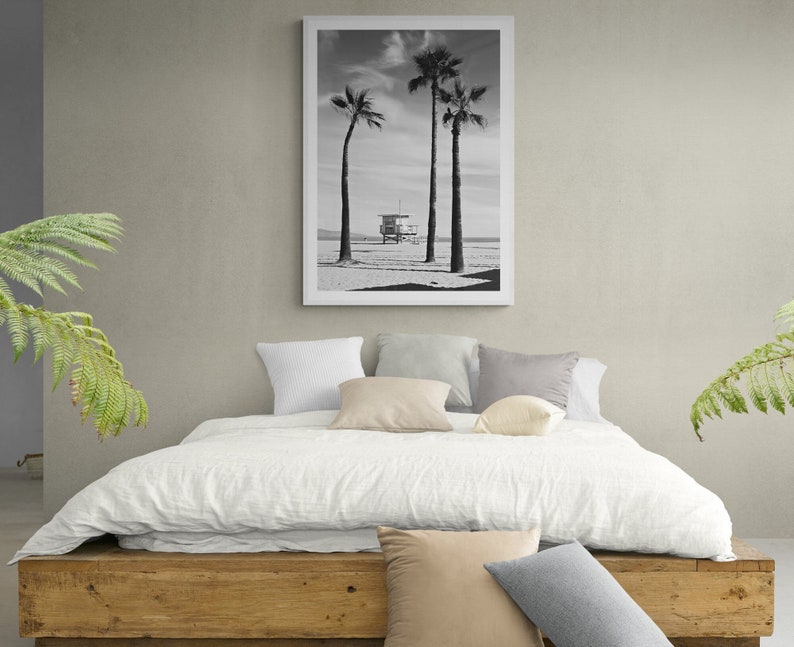 Puede incluir: Una fotograf&iacute;a en blanco y negro de tres palmeras en una playa con una caseta de salvavidas en el fondo. La fotograf&iacute;a est&aacute; enmarcada y colgada en una pared sobre una cama blanca con almohadas.