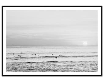 May include: A black and white photograph of a seascape with surfers in the water. The horizon is in the distance, and the sky is clear. The sun is setting in the distance, casting a warm glow on the water.