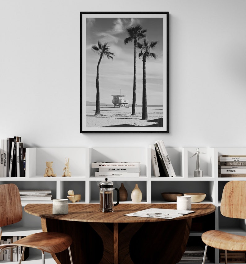 May include: A black and white photograph of three palm trees on a beach with a lifeguard stand in the distance. The photograph is framed in a black frame and is hanging on a white wall above a white bookshelf with books and decorative objects.