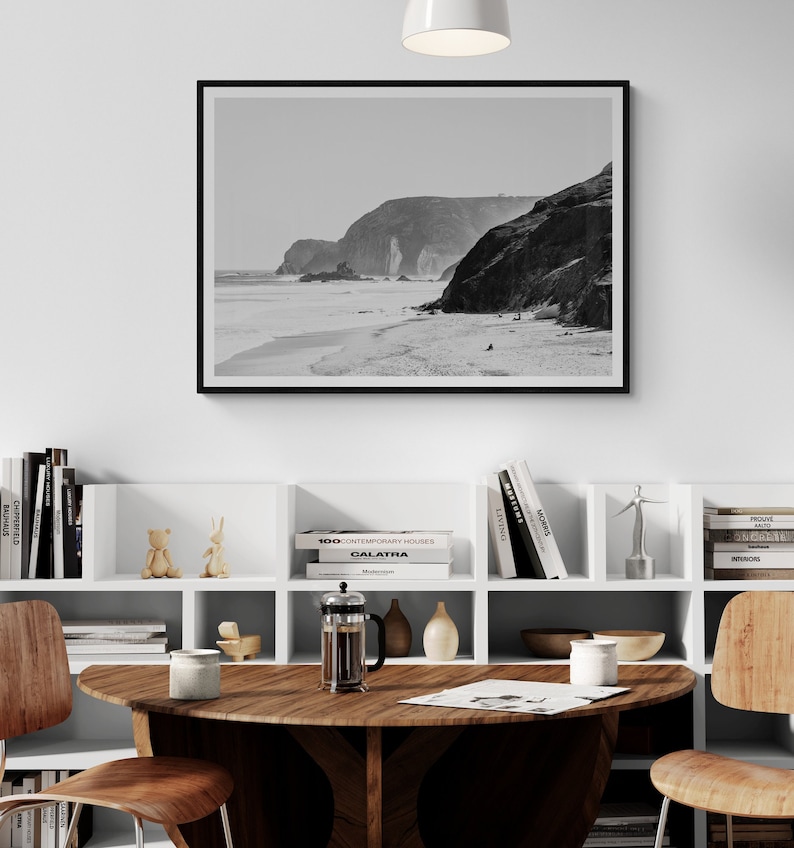 May include: A black and white photograph of a rocky coastline with a beach in the foreground. The photo is framed in a black frame and hanging on a white wall above a white bookshelf.