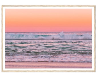 Può includere: Un tramonto rosa e arancione su un oceano turchese con onde bianche e spumose che si infrangono sulla riva. La sabbia è di colore rosa chiaro.