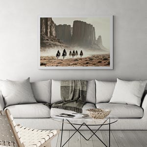 May include: A framed photograph of a group of people riding horses through a desert landscape. The photograph is in a white frame and features a muted colour palette of browns, tans, and greys. The horses are moving in a line, and the riders are wearing dark clothing.