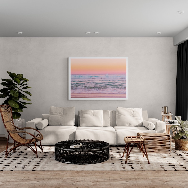 May include: A white sofa with pillows sits in a living room with a white rug and a black woven coffee table. A framed photograph of a beach scene with pink and blue hues hangs on the wall above the sofa.