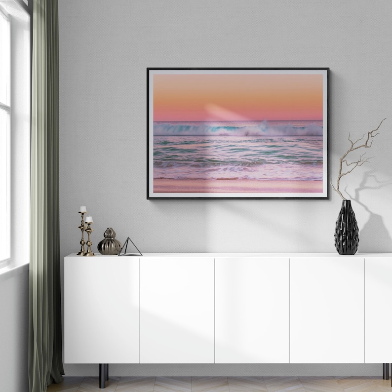 May include: A framed photograph of a pink and blue sunset over a beach with waves crashing on the shore. The photo is hanging on a white wall above a white cabinet with black legs.