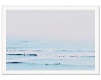 May include: A photograph of a seascape with surfers in the water. The water is a light blue color and the sky is a pale blue. The surfers are silhouetted against the water and sky.