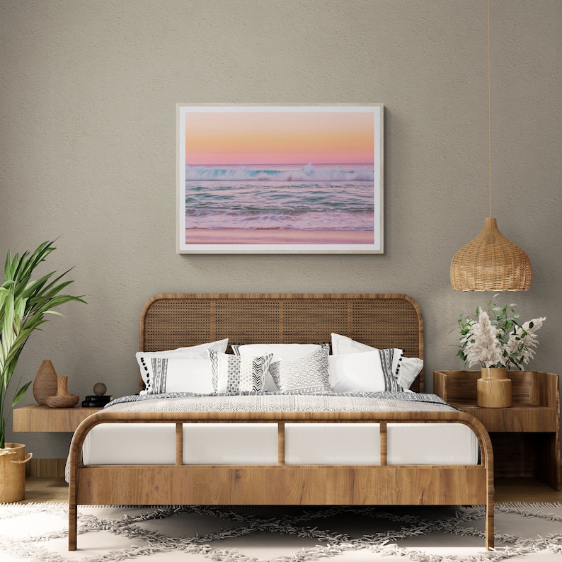 May include: A wooden bed frame with a woven headboard and a white bedspread. The bed is decorated with white and black patterned pillows. A framed photograph of a beach scene with a pink and blue sunset hangs on the wall above the bed.