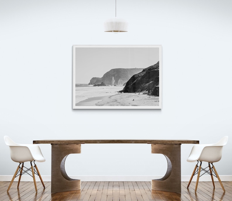 May include: A black and white photograph of a beach scene with a rocky cliff in the background. The photo is framed and hanging on a white wall above a wooden table with two white chairs.