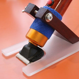 May include: A grooming tool featuring a wooden handle, a gold-coloured body, and a black head with a white blade. It is supported by a clear plastic base and has a blue rubber grip. The tool is designed for grooming.