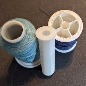 Puede incluir: Tres carretes de hilo, uno azul, uno azul oscuro y uno blanco. Los carretes están sobre una superficie gris oscuro.