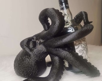 以下が含まれることがあります： 黒いタコの形をしたボトルホルダーに、クラーケンラムのボトルが入っています。タコは表面がざらざらしていて、触手でボトルを保持しています。