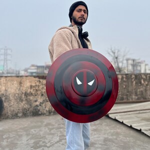 Puede incluir: Una persona sosteniendo un escudo de Capit&aacute;n Am&eacute;rica rojo, negro y blanco con un dise&ntilde;o de m&aacute;scara de Deadpool.