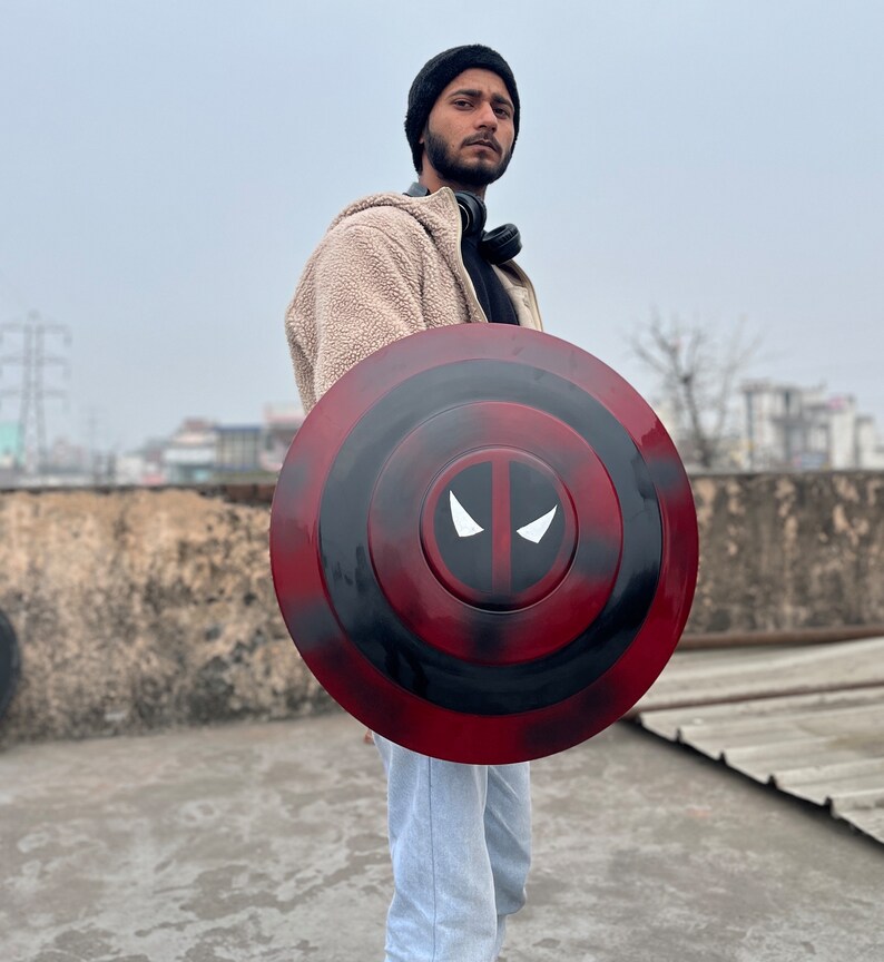 Puede incluir: Una persona sosteniendo un escudo de Capit&aacute;n Am&eacute;rica rojo, negro y blanco con un dise&ntilde;o de m&aacute;scara de Deadpool.