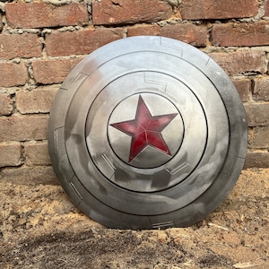 May include: A silver-colored, circular shield with a red star in the center. The shield has a weathered appearance and is divided into segments. The background is a weathered brick wall.