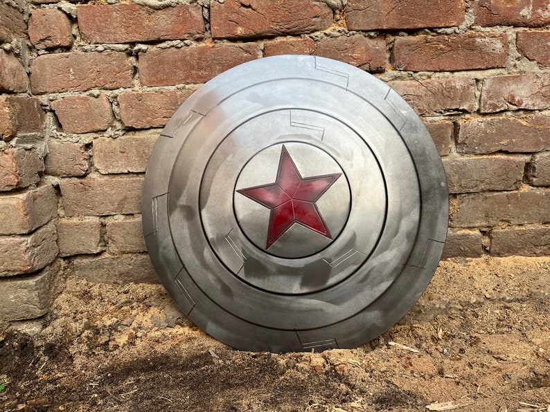 May include: A silver-colored, circular shield with a red star in the center. The shield has a weathered appearance and is divided into segments. The background is a weathered brick wall.