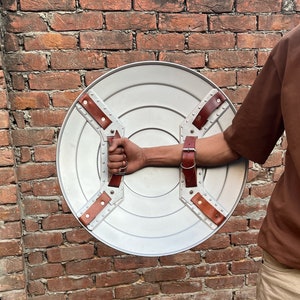 May include: A round, silver-colored shield with riveted metal supports and brown leather straps. The shield has a handle and an arm strap for secure grip. The background is a weathered brick wall.