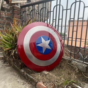 May include: A red, white, and blue Captain America shield with a silver star in the center. The shield is lying on the ground in front of a brick wall.