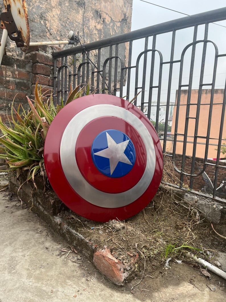 May include: A red, white, and blue Captain America shield with a silver star in the center. The shield is lying on the ground in front of a brick wall.