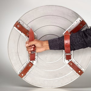 Puede incluir: Un escudo de Capit&aacute;n Am&eacute;rica plateado y rojo con correas de cuero marr&oacute;n. El escudo est&aacute; sostenido por una mano.