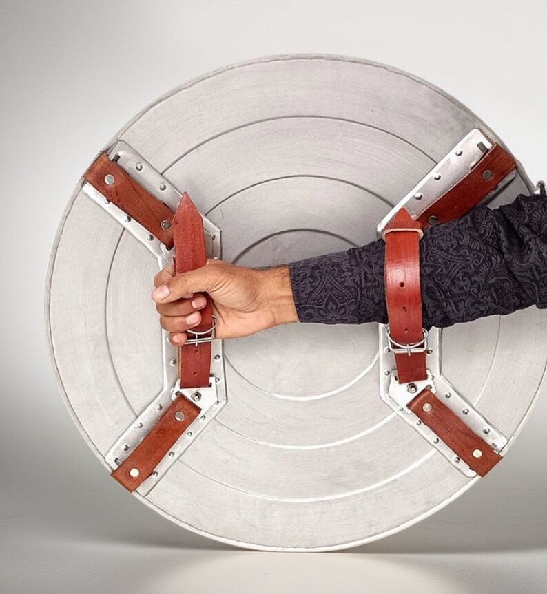 Puede incluir: Un escudo de Capit&aacute;n Am&eacute;rica plateado y rojo con correas de cuero marr&oacute;n. El escudo est&aacute; sostenido por una mano.