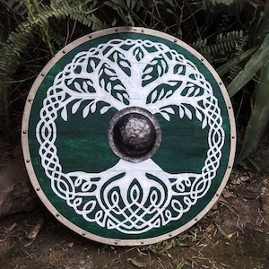 May include: A round wooden shield featuring a green background and a white Celtic tree of life design. The shield has a metal boss in the centre and is trimmed with metal studs. The shield is approximately 76 cm in diameter.
