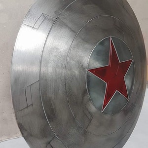 May include: A metallic gray shield with a red star emblem at the center. The shield has a circular design with concentric rings and rectangular patterns. The star is a vibrant red with a silver outline.