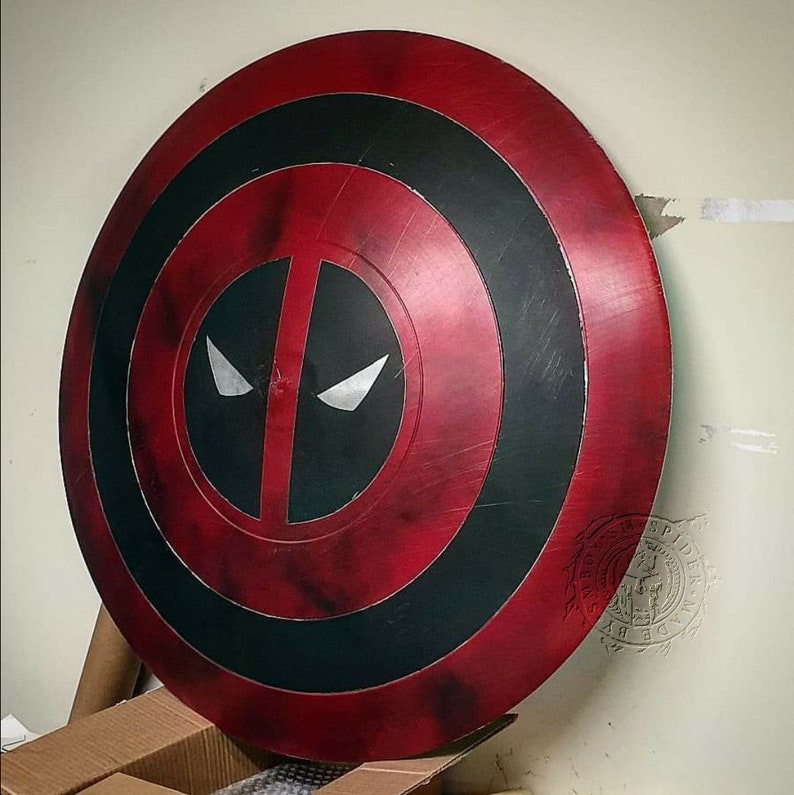 Puede incluir: Un escudo de Capit&aacute;n Am&eacute;rica rojo y negro con un dise&ntilde;o de m&aacute;scara de Deadpool. El escudo tiene un aspecto desgastado con un efecto de salpicadura de pintura roja y negra.