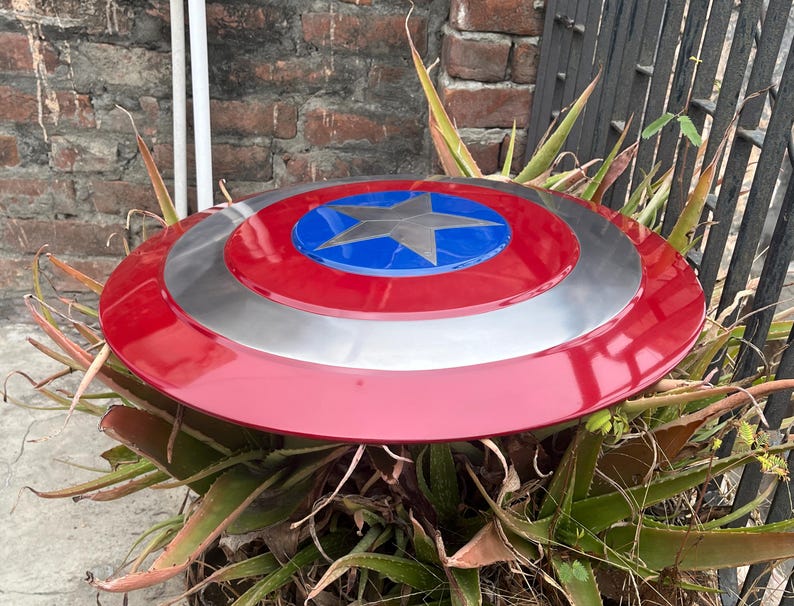 May include: A red, white, and blue Captain America shield with a silver metal rim. The shield is sitting on top of a green plant.