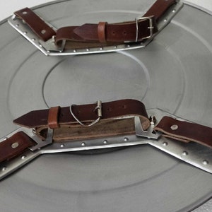 Puede incluir: Un escudo de metal con tres correas de cuero marr&oacute;n unidas a la parte posterior. Las correas tienen hebillas met&aacute;licas y est&aacute;n dise&ntilde;adas para ser usadas en la espalda.