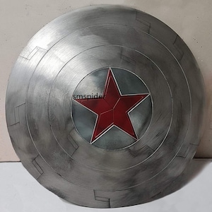 May include: A silver-colored, circular shield with a red star emblem at the center. The shield has a distressed, metallic finish with concentric circular patterns and rectangular details. The star is a vibrant red.