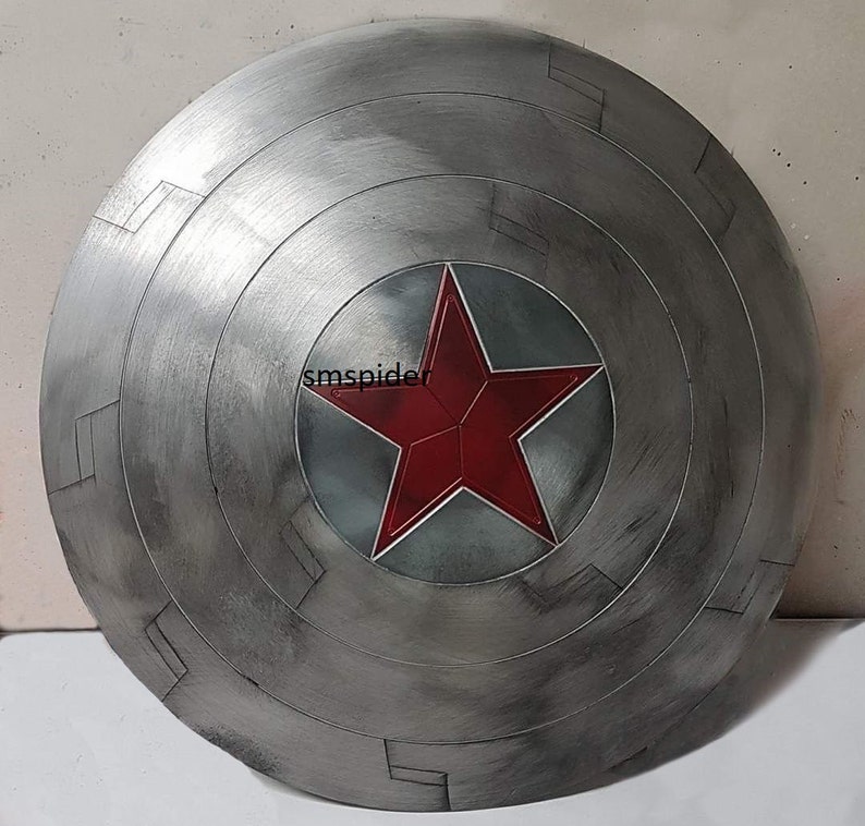 May include: A silver-colored, circular shield with a red star emblem at the center. The shield has a distressed, metallic finish with concentric circular patterns and rectangular details. The star is a vibrant red.