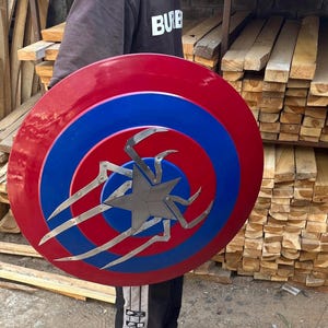 Puede incluir: Un escudo de Capit&aacute;n Am&eacute;rica rojo, blanco y azul con una estrella plateada y un dise&ntilde;o de ara&ntilde;a en el centro.