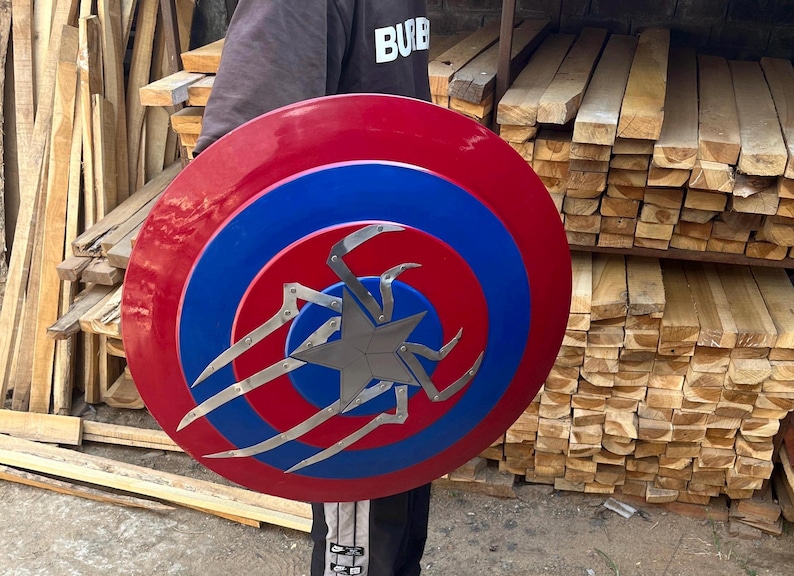 Puede incluir: Un escudo de Capit&aacute;n Am&eacute;rica rojo, blanco y azul con una estrella plateada y un dise&ntilde;o de ara&ntilde;a en el centro.