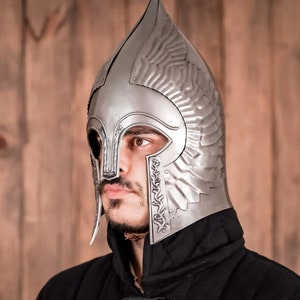 May include: A silver helmet with a pointed top and decorative wings. The helmet has a face mask with a small opening for the eyes. The helmet is worn by a person with a beard.