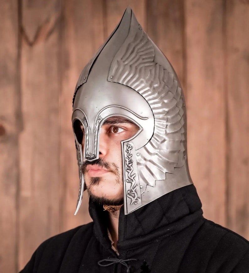 May include: A silver helmet with a pointed top and decorative wings. The helmet has a face mask with a small opening for the eyes. The helmet is worn by a person with a beard.