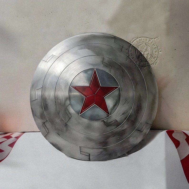 May include: A silver-colored, circular shield with a prominent red star at its center. The shield has concentric rings and rectangular cutouts, with a weathered, metallic appearance. The star is outlined in silver.