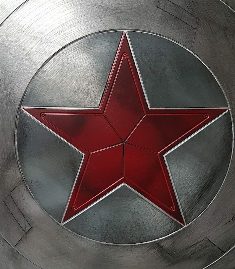 May include: A close-up of a metallic shield with a large, red star in the center. The star is outlined in a lighter red and set against a gray circular background. The shield has a brushed metal finish.