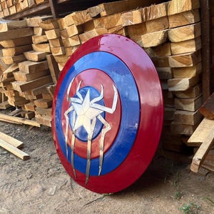 Puede incluir: Un escudo de Capit&aacute;n Am&eacute;rica rojo, blanco y azul con un dise&ntilde;o de ara&ntilde;a plateado en el centro.