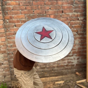 May include: A silver-colored shield with a red star in the center. The shield has concentric circles and rectangular details. The shield is held in front of a person against a brick wall background.