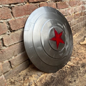 May include: A metallic silver shield with a red star emblem in the center. The shield has a circular design with concentric rings and rectangular details. It leans against a weathered brick wall, suggesting a superhero theme.