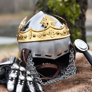 May include: A medieval helmet with a gold crown and chainmail. The helmet is made of metal and has a pointed top. The chainmail is made of metal rings linked together.