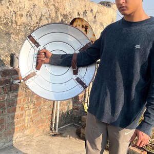 May include: A person holding a large, round, silver shield with brown leather straps. The shield has a raised, circular design in the center.