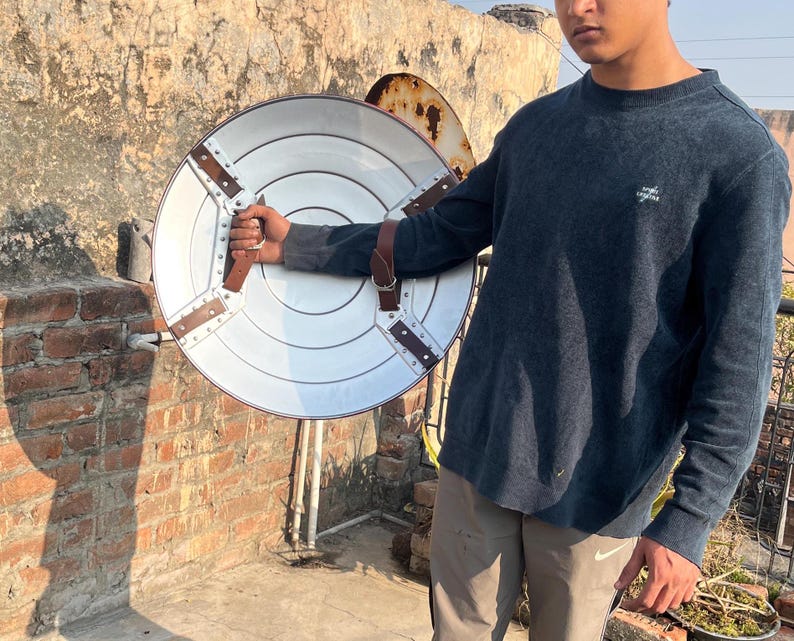 May include: A person holding a large, round, silver shield with brown leather straps. The shield has a raised, circular design in the center.