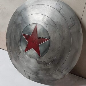 May include: A silver-colored, circular shield with a red star emblem at the center. The shield has a weathered appearance with concentric circular patterns and rectangular details. The shield is a prop or collectible.