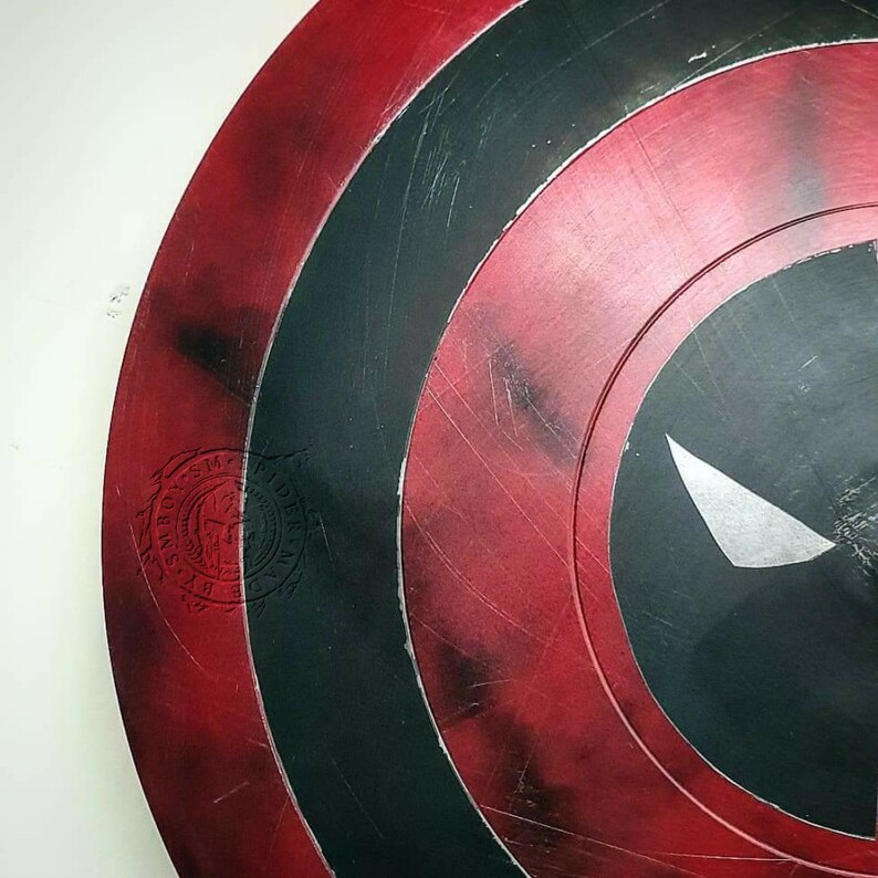 Puede incluir: Un escudo de Capit&aacute;n Am&eacute;rica rojo, blanco y negro con una calavera negra y ojos blancos pintados en el centro. El escudo tiene un aspecto desgastado con ara&ntilde;azos y astillas de pintura. El escudo tiene un sello circular en el lateral que dice "SM BOX SM" con una calavera y huesos cruzados en el centro.