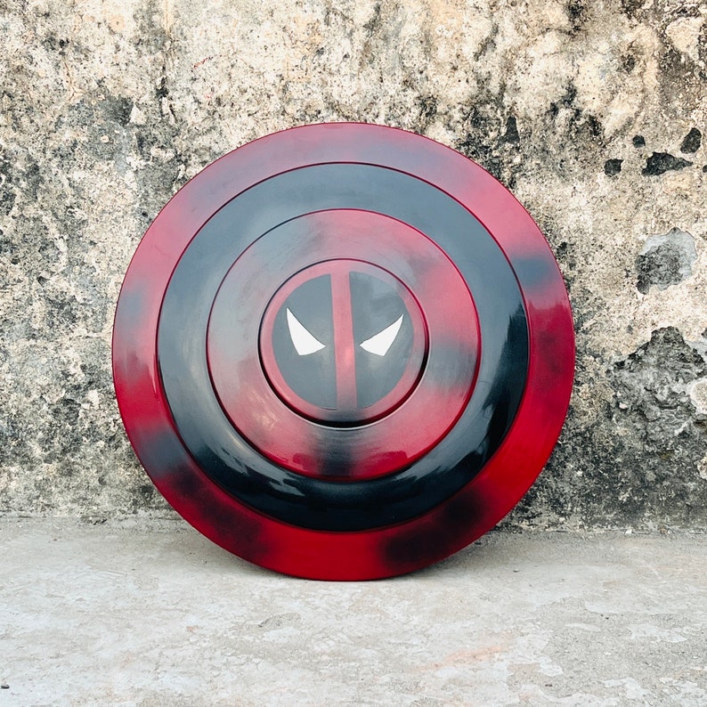 Puede incluir: Un escudo circular rojo y negro con un logotipo de Deadpool blanco en el centro. El escudo tiene un dise&ntilde;o de c&iacute;rculos conc&eacute;ntricos con un anillo exterior negro y un anillo interior rojo.