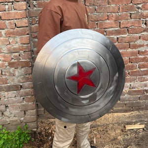 May include: A silver-colored, circular shield with a red star in the center. The shield has a weathered appearance with concentric circles and rectangular details. The shield is held in front of a person.