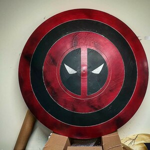 Puede incluir: Un escudo de Capit&aacute;n Am&eacute;rica rojo, blanco y negro con una m&aacute;scara de Deadpool pintada en la parte delantera. El escudo tiene un aspecto desgastado, con pinceladas y salpicaduras de pintura visibles.